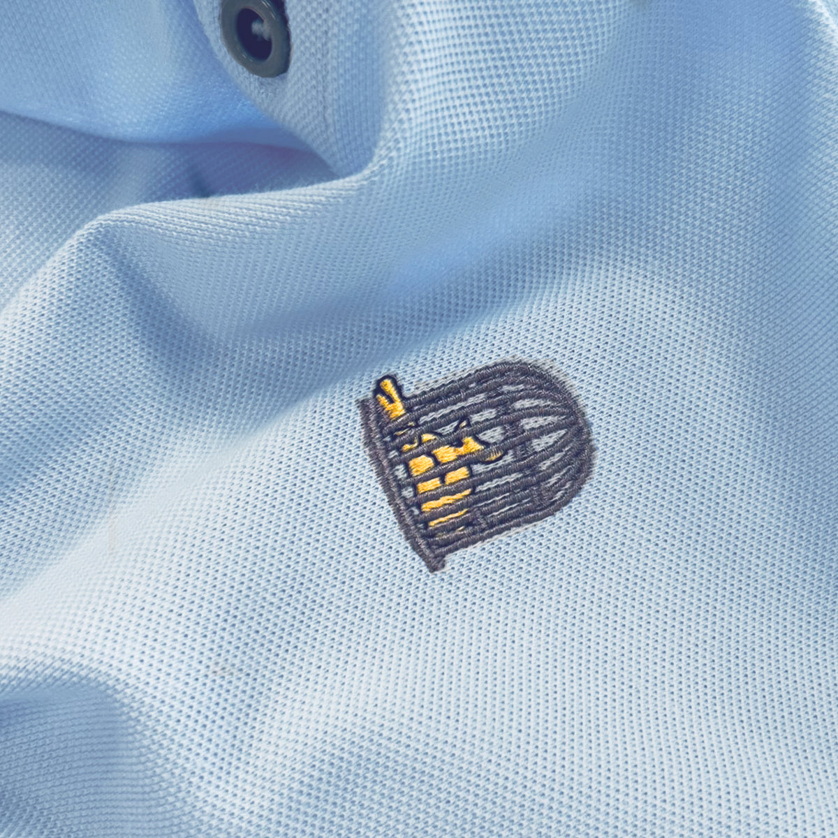 Close-up detail of the embroidered Sauce & Brown birdcage logo on the Calma light blue polo shirt fabric.