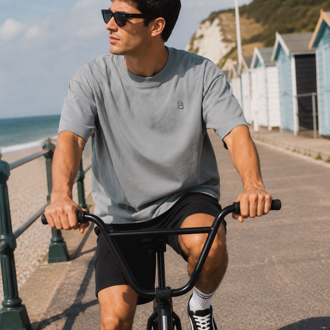 Male model wearing the Sauce & Brown Haze oversized t-shirt in light grey while riding a BMX cruiser along a coastal promenade with beach huts in the background.