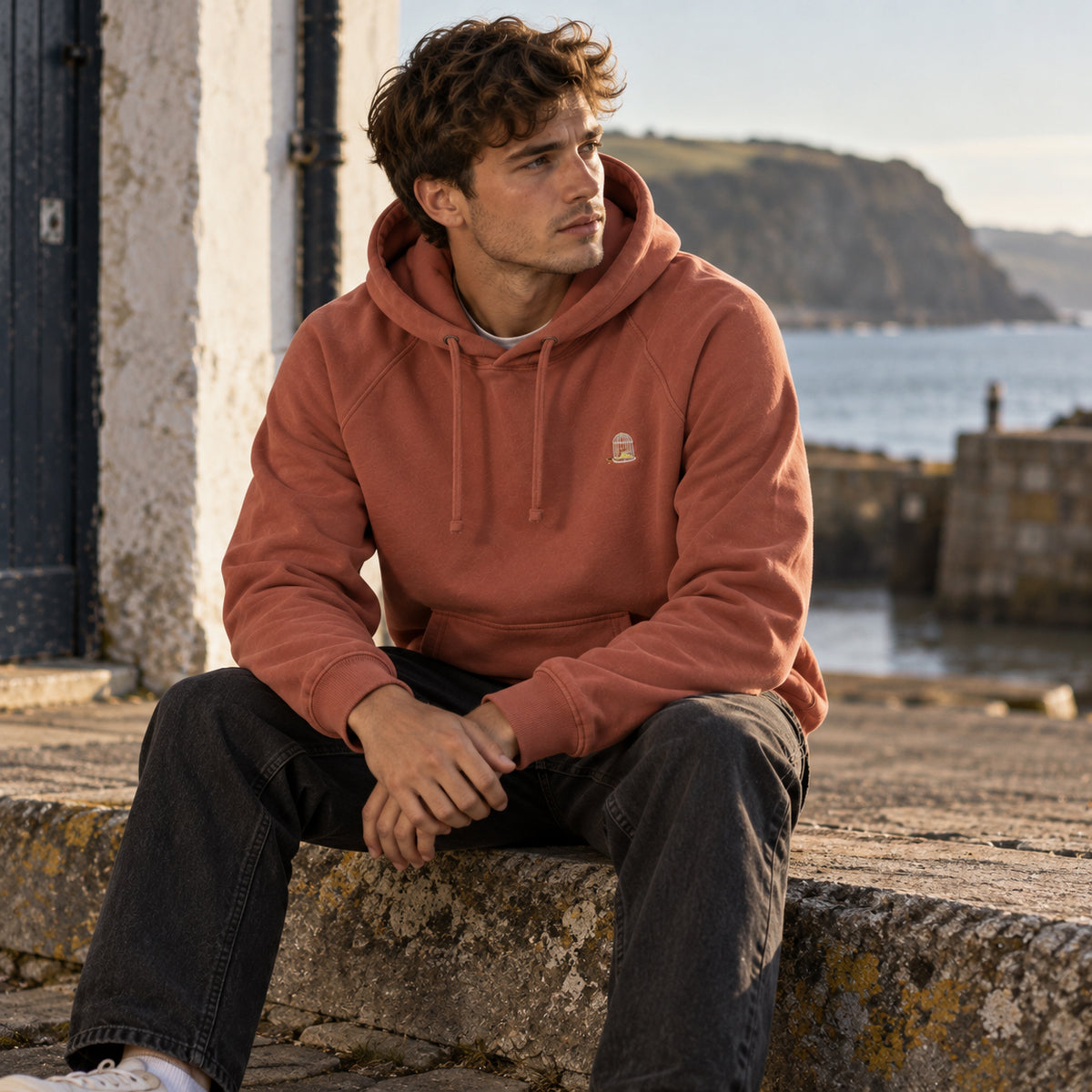 Man wearing the Sauce & Brown Kiln Hoody in Vintage Washed Terracotta, seated by a coastal harbour. Heavyweight 400gsm organic cotton with a relaxed fit.