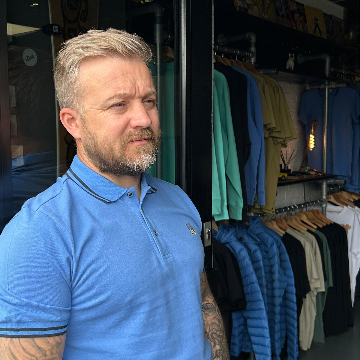 Man wearing blue Mariner polo shirt standing in shop doorway with clothing racks behind